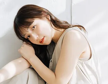 白い壁際で首をかしげる茶髪の女性